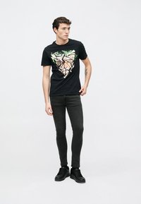 Sort bomuld t-shirt med et geometrisk grafisk design i nuancer af grøn, pink og gul, kombineret med sorte skinny jeans og sneakers.