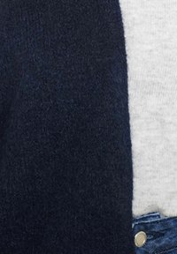 Tumši zilas krāsas biezs adīts kardigāns ar mīkstu tekstūru, apvienots ar gaiši pelēku T-kreklu un džinsu biksēm, izceļot metāla pogas detaļu.