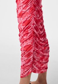 Roza in rdeča midi krilo z rožnatim vzorcem z nabrano detajlo po sredini, nošeno s črnimi čevlji z odprtimi prsti.