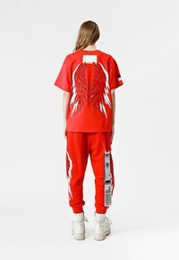 Camisa corta roja de mangas y joggers a juego que presentan diseños de alas en blanco y rojo. Detalle de la etiqueta en los pantalones. El modelo lleva botas blancas.