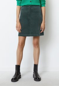 Dunkelgrüder Cord-Rock mit geradem Schnitt, vorderem Knopfverschluss und subtiler Textur. Kombiniert mit schwarzen Stiefeletten.