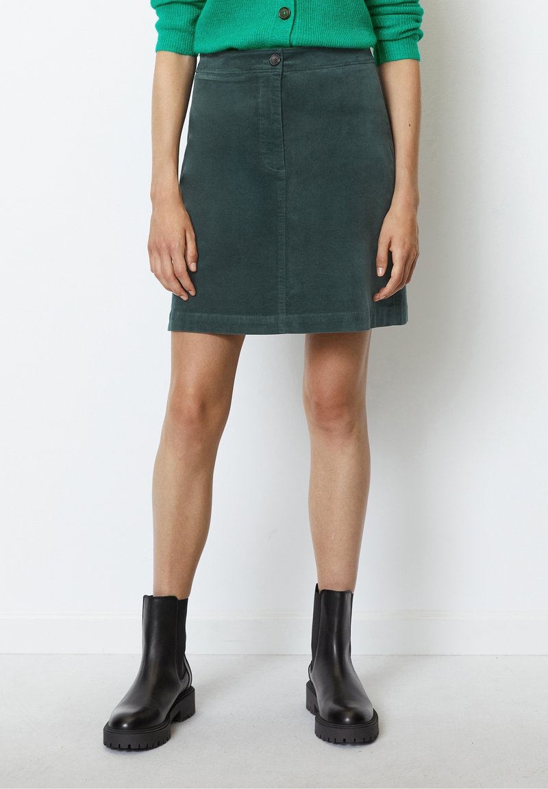 Dunkelgrüder Cord-Rock mit geradem Schnitt, vorderem Knopfverschluss und subtiler Textur. Kombiniert mit schwarzen Stiefeletten.