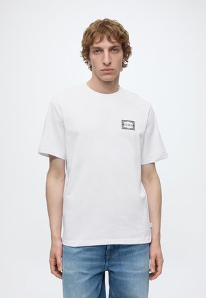 Jeune homme aux cheveux bouclés châtain clair, portant un t-shirt blanc à manches courtes avec un petit écusson rectangulaire et un jean bleu, debout face à l'avant.