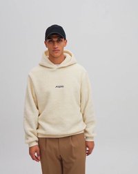 Cremefarbene Fleece-Hoodie mit Kordelzugkapuze und kleinem schwarzem Logo auf der Brust, kombiniert mit beigen Hosen und einer schwarzen Kappe.