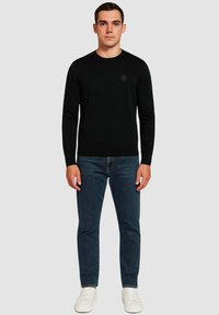 Maglione nero a maglia con scollo rotondo, abbinato a jeans di denim scuro e sneakers bianche; in piedi contro uno sfondo neutro.