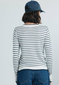 Maglione a righe bianco con strisce blu navy, collo e polsini a coste. Abbinato a pantaloni di jeans blu. Presenta dettagli con bottoni sulle maniche.