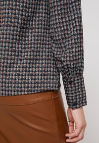Camicia a quadrati marrone e blu con maniche lunghe, polsini abbottonati e dettagli plissettati, abbinata a una gonna in pelle marrone.