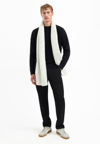 Schwarzer Strickpullover, hellgrauer gerippter Schal, schwarze Hose und graue Sneaker mit weißen Streifen. Model steht vor einem weißen Hintergrund.