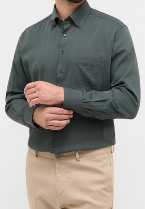 Homme portant une chemise boutonnée vert foncé et un pantalon beige ajustant le poignet gauche de sa chemise avec sa main droite.