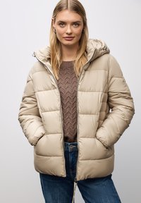 Giacca puffer beige con cappuccio, caratterizzata da un design trapuntato, chiusura frontale con zip e due tasche laterali. Indossata sopra un maglione marrone a trama.