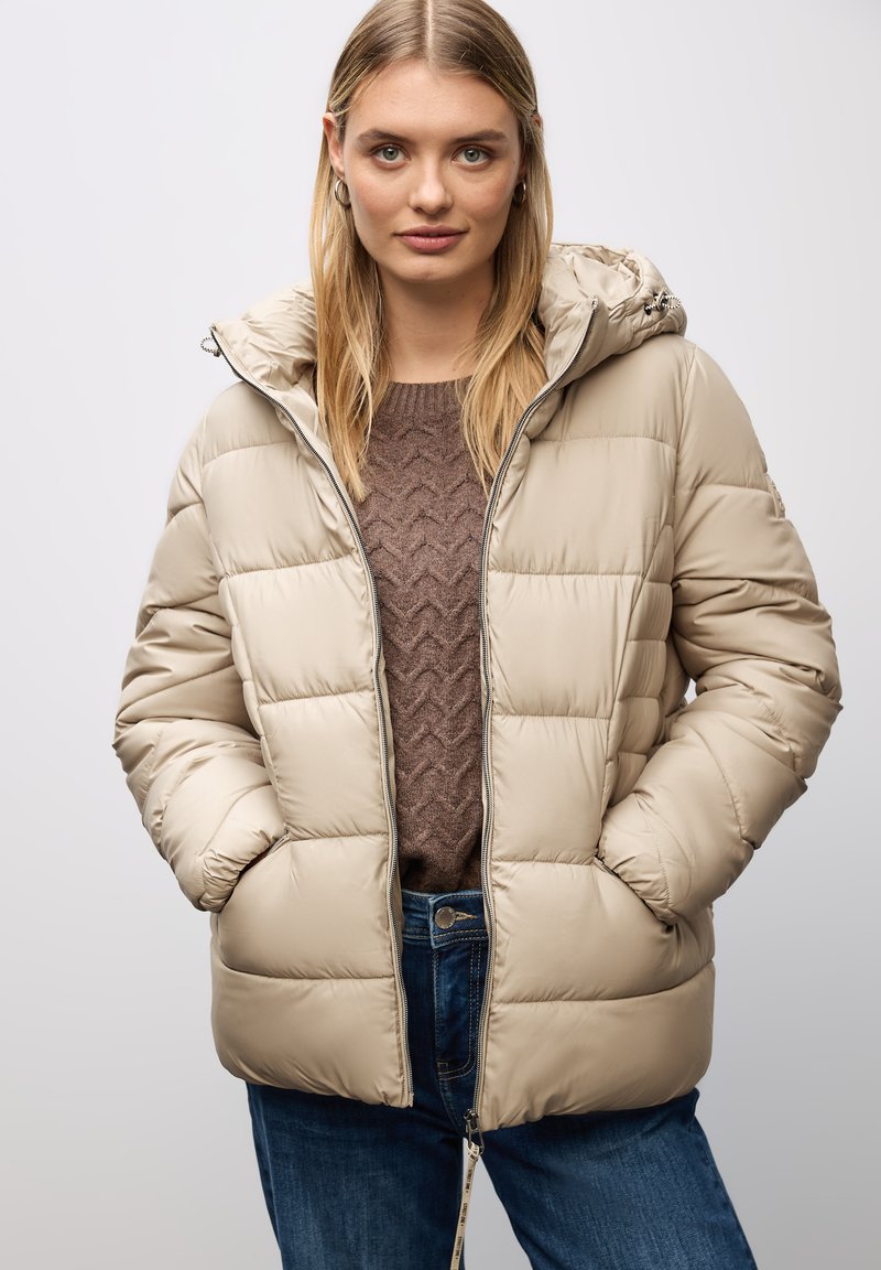 Giacca puffer beige con cappuccio, caratterizzata da un design trapuntato, chiusura frontale con zip e due tasche laterali. Indossata sopra un maglione marrone a trama.