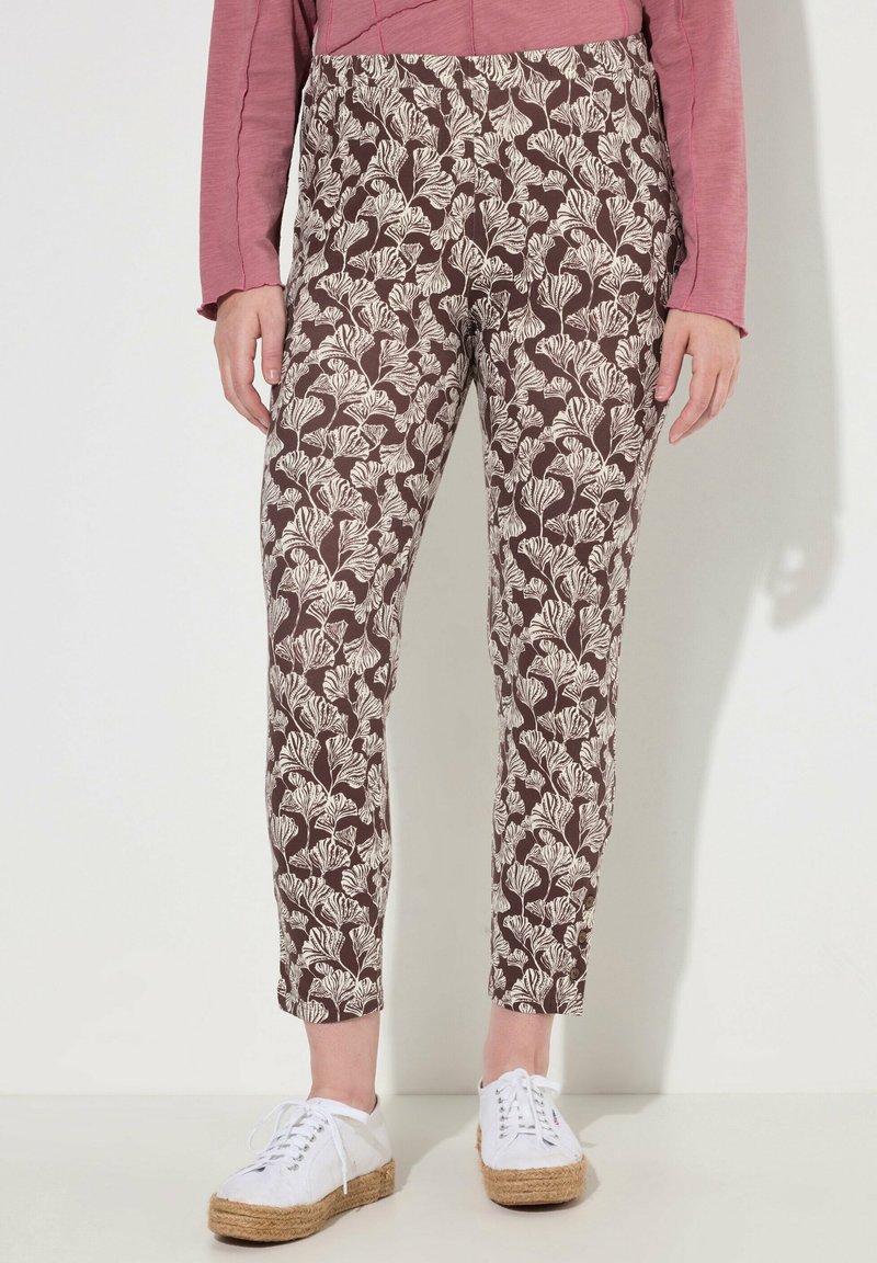 Pantaloni marroni a fantasia con design floreale bianco, aderenti in vita e affusolati alla caviglia, abbinati a scarpe bianche con lacci.