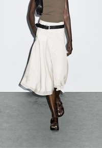 Jupe midi blanche, fluide, avec un large ourlet, associée à une ceinture noire et des sandales brunes. Texture lisse, design décontracté et minimalisme des détails.