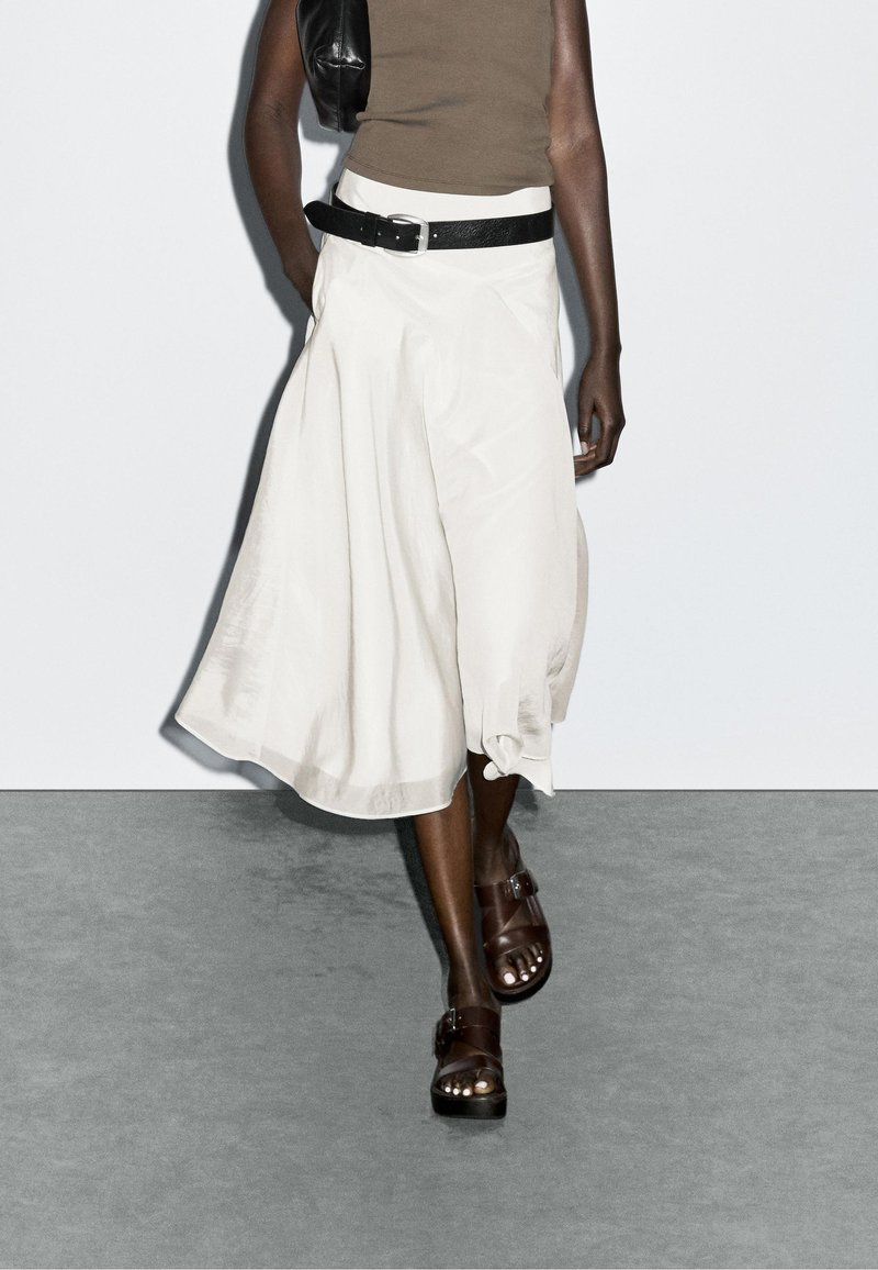 Jupe midi blanche, fluide, avec un large ourlet, associée à une ceinture noire et des sandales brunes. Texture lisse, design décontracté et minimalisme des détails.