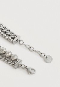 Bracciale in tonalità argento con diversi stili di catena, perline rotonde, chiusura a gancio e charm circolare brandizzato su una catena di estensione.