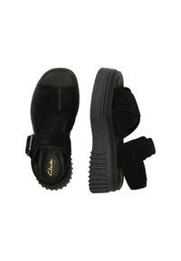 Mustat platform-sandaalit, joissa on mokkanahkainen ristikkäisnauha, säädettävä nilkkahihna, teksturoitu paksu pohja ja "Clarks Signature" -merkintä jalkapohjassa.