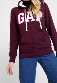 Femme portant un sweat à capuche zippé GAP bordeaux avec des cordons blancs et un jean bleu, visible des épaules aux hanches sur un fond uni.