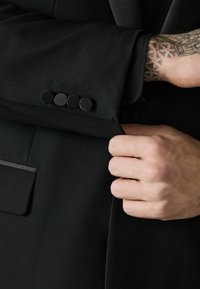 Hand stellt die Manschette eines schwarzen Sakkos mit vier schwarzen Knöpfen ein, sichtbares Tattoo am Handgelenk und Unterarm.