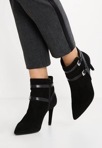 Zwarte suède enkelboots met puntige neuzen, voorzien van zwarte leren straps en zilveren gespen, geplaatst op slanke hoge hakken.