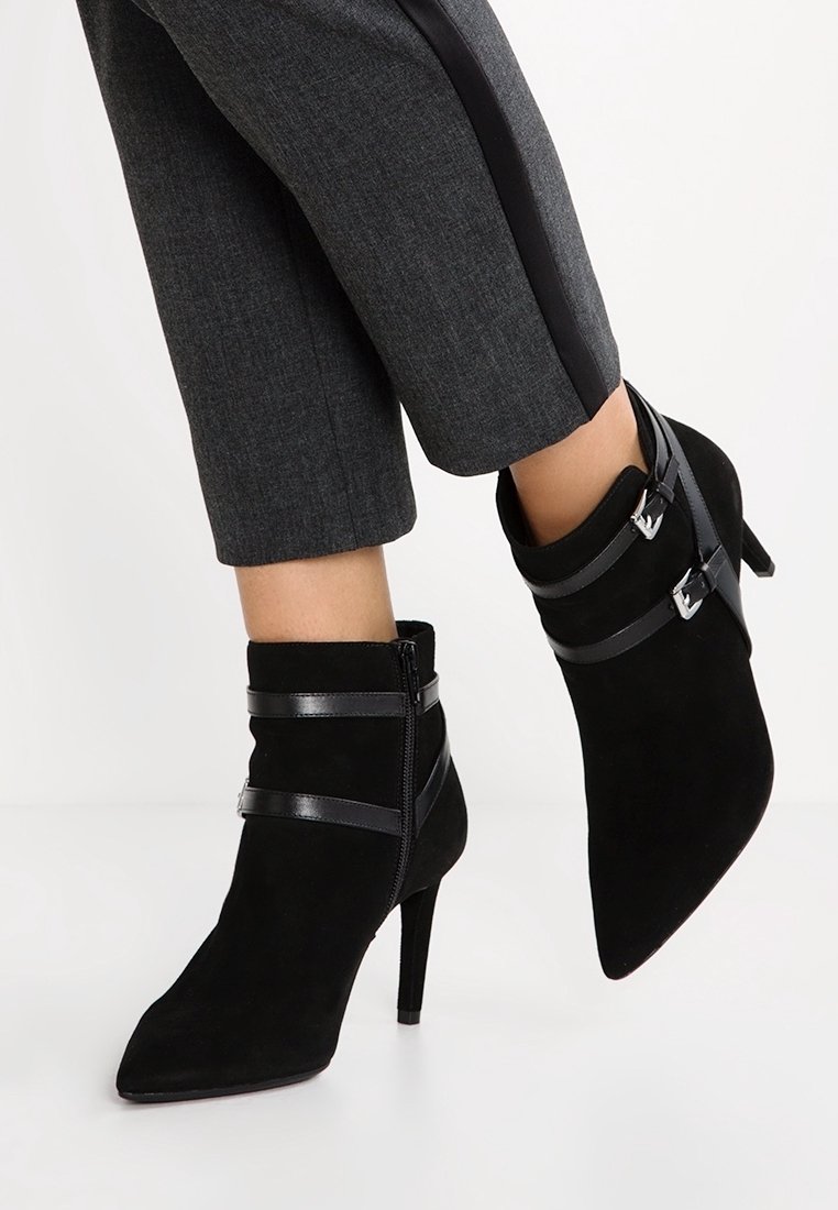 Bottines en suede noir avec des pointes pointues, dotées de sangles en cuir noir et de boucles en argent, reposant sur des talons hauts fins.