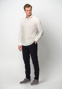 Camicia botton down beige chiaro con maniche lunghe, abbinata a pantaloni blu scuro e sneakers grigie. Tessuto morbido, vestibilità adattata, design minimalista.