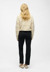 Blonďatá žena stojí zády, má na sobě krémový svetr s copánkovým vzorem, černé rovné kalhoty a béžové ploché boty proti jednolitému pozadí.