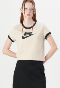 Beige zkrácený tričko s černým výstřihem a manžetami, na přední straně s výrazným černým logem Nike. Měkká tkanina se volným střihem.