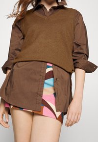 Chemise en coton marron à boutons, portée sous un gilet sans manches en maille marron, associée à une mini-jupe à motifs colorés aux nuances de bleu, rose et marron.