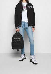 Černá univerzitní bunda s bílými pruhy, bílé tričko s potiskem, světle modré džíny a černý batoh Versace Jeans Couture. Bílé tenisky.