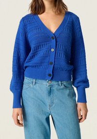 Blå stickad kofta med djup V-ringning, strukturerat mönster och fem framknappar. Långa ärmar med ribbade manschetter och midja.