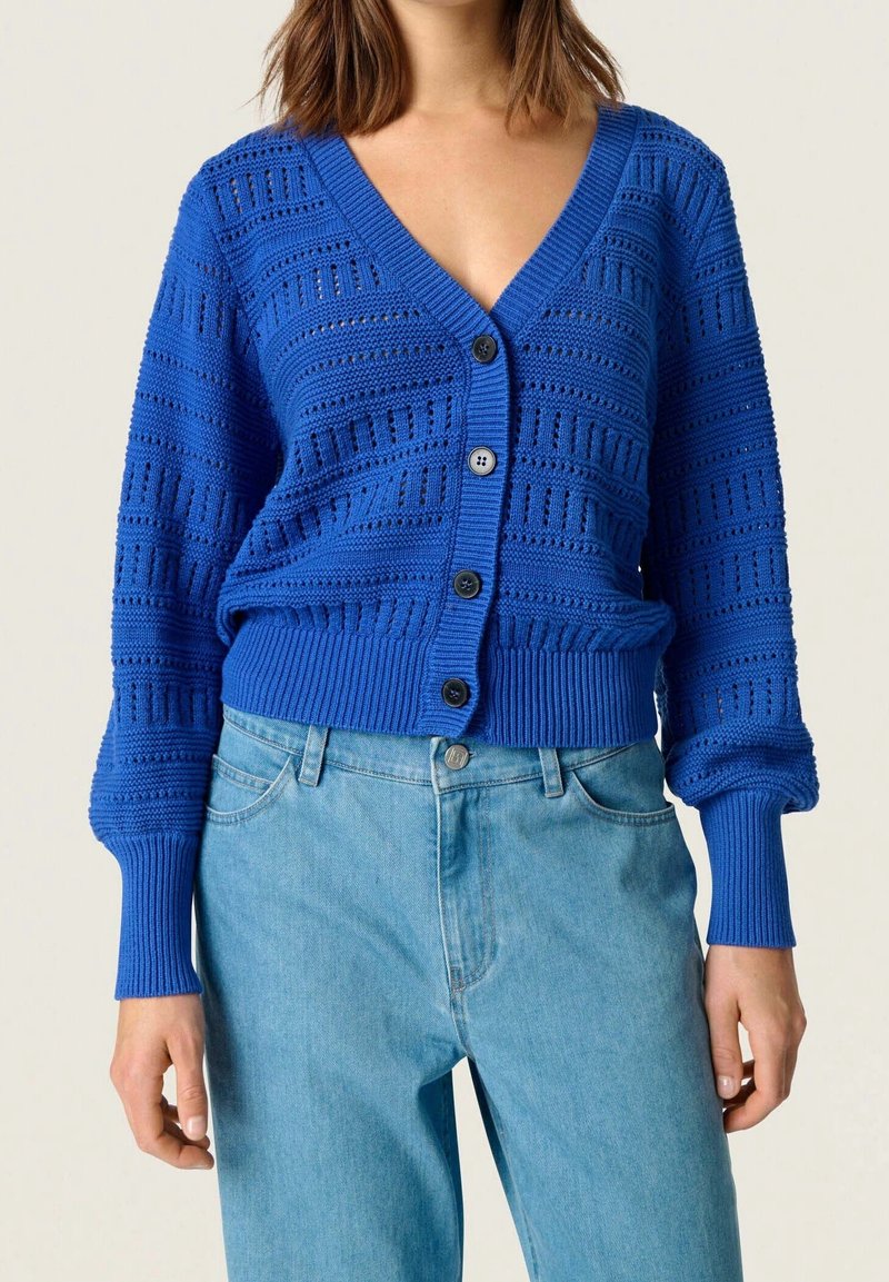 Blå stickad kofta med djup V-ringning, strukturerat mönster och fem framknappar. Långa ärmar med ribbade manschetter och midja.