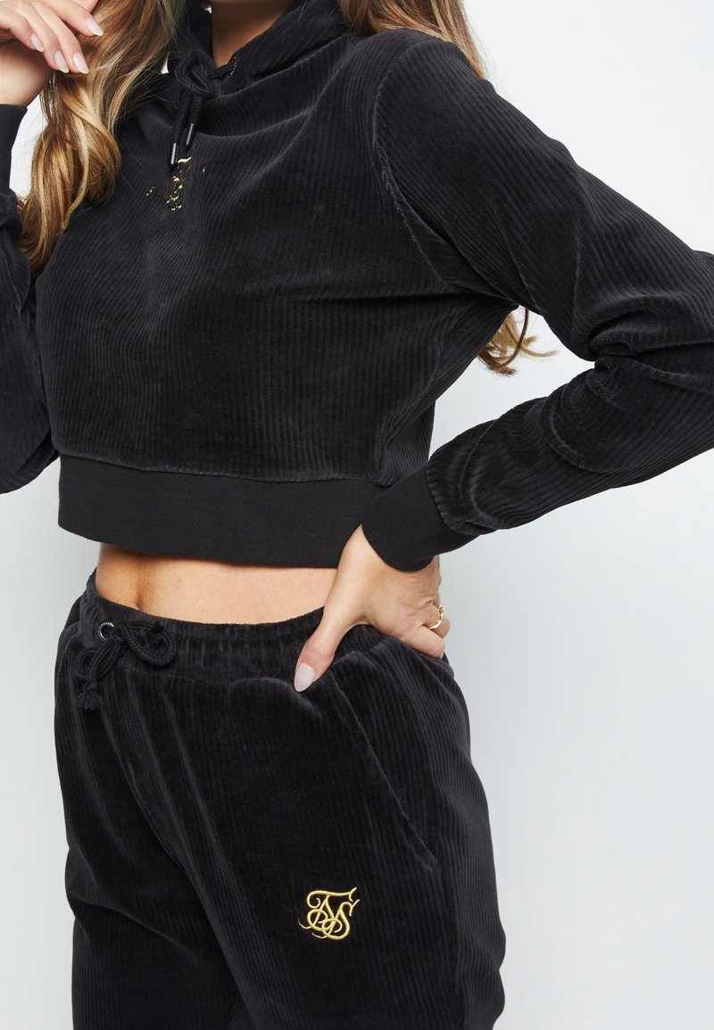 Schwarzes Cord-Crop-Top mit hohem Kragen und verstellbarem Kordelzug. Kombiniert mit passenden Kordelhosen mit goldenem, gesticktem Logo.