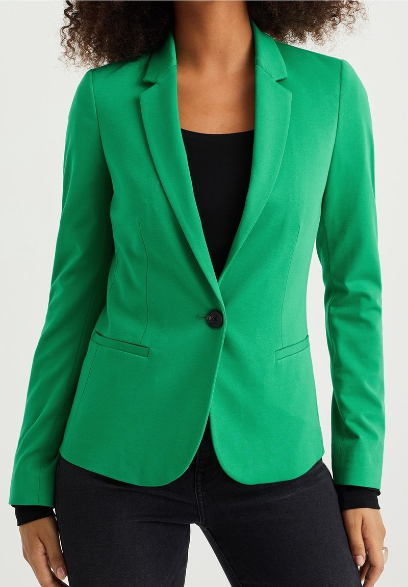 Grön blazer i smidig tyg, med figurnära design och enkel knappstängning, samt framfickor, matchad med en svart topp och mörka jeans.