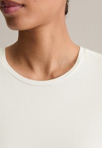 Nahaufnahme des Nackens und des unteren Gesichts einer Person, die ein einfaches weißes Rundhals-T-Shirt vor einem neutralen Hintergrund trägt.