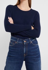 Pull bleu marine à manches longues en tissu tricoté, associé à un jean taille haute bleu foncé avec des accessoires et des coutures en ton cuivré.