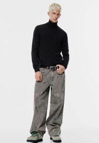 Črn pulover s potiskom, sivi široki jeans z teksturiranim vzorcem in čevlji z zelenimi vezalkami. Model stoji z rokami na pasu.