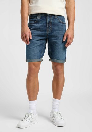 Shorts in denim blu medio con orlo arrotolato, dotati di cinque tasche e zip. Abbinati a scarpe da ginnastica bianche e calzettoni bianchi.