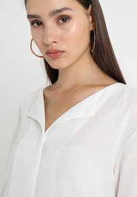 White, lightweight blouse with a v-neckline and collar detail, smooth texture, and long sleeves; model wearing large hoop earrings.
