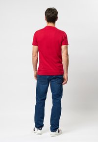 Man standing facing away, wearing a red t-shirt, blue jeans, and white sneakers with navy and red stripes.