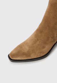 Brūnie zamša potītes zābaki ar smailu purngalu, gludu tekstūru un melnu zoli. Piedāvā elegantus dizainus un minimālus šuvju akcentus.
