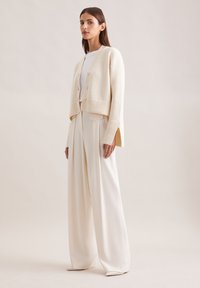Crème geknipt cardigan met geribbelde manchetten over een witte top, gecombineerd met brede crème broek. Gladde textuur, eenvoudige knoopdetails.