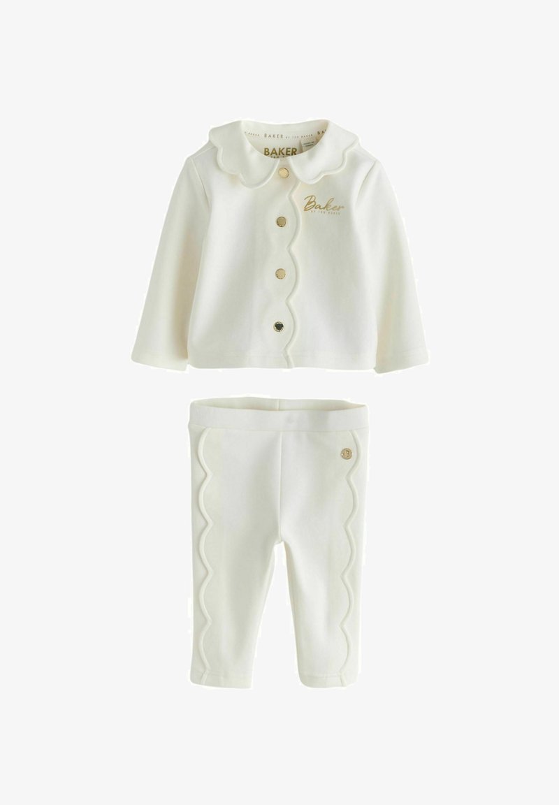 Dviejų dalių kūdikio apranga kreminės spalvos. Viršutinė dalis turi banguotą apykaklę ir auksinius mygtukus; kelnės puoštos banguotu apvadu ir auksiniu emblematu. Minkšta medžiaga.