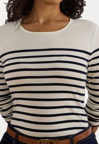 Maglietta a maniche lunghe con base crema e righe orizzontali blu scuro. Tessuto morbido e scollo rotondo. Indossata con una cintura marrone.
