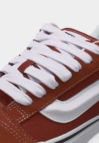 Gros plan sur une chaussure de sport rouge rouille avec une bande en cuir blanc et des lacets épais blancs passés dans des œillets en métal.