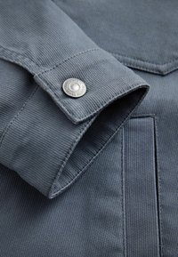 Poignet de manche en tissu gris avec un bouton-pression métallique et une couture de poche visible sur une chemise ou une veste texturée.