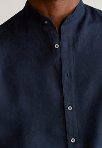 Camicia di lino blu navy con colletto a stand, dotata di bottoni in argento e una texture liscia. Design minimalista con patta nascosta.