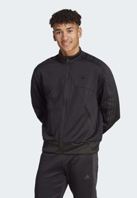 Chándal negro de Adidas con chaqueta con cremallera y pantalones a juego. Presenta tres rayas en las mangas, tela suave y puños acanalados.