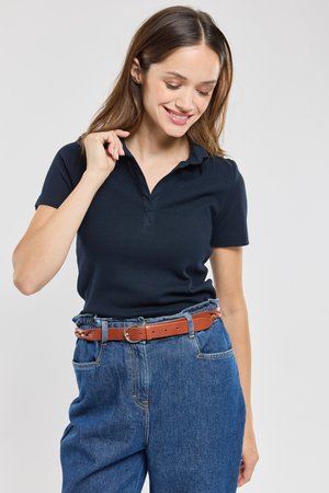 Kvinde smilende, iført en navyblå kortærmet polo, stukket ned i højtaljede blå jeans med et brunt bælte.