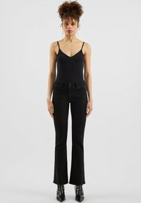 Schwarze, ausgestellte Jeans mit schmaler Taille, aus Denim gefertigt. Kombiniert mit einem schwarzen ärmellosen Bodysuit. Glänzende, spitze Stiefeletten.