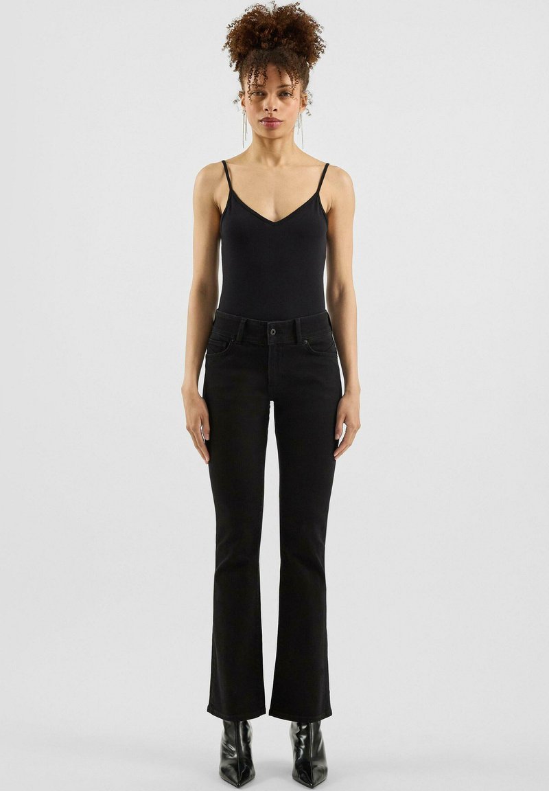 Schwarze, ausgestellte Jeans mit schmaler Taille, aus Denim gefertigt. Kombiniert mit einem schwarzen ärmellosen Bodysuit. Glänzende, spitze Stiefeletten.